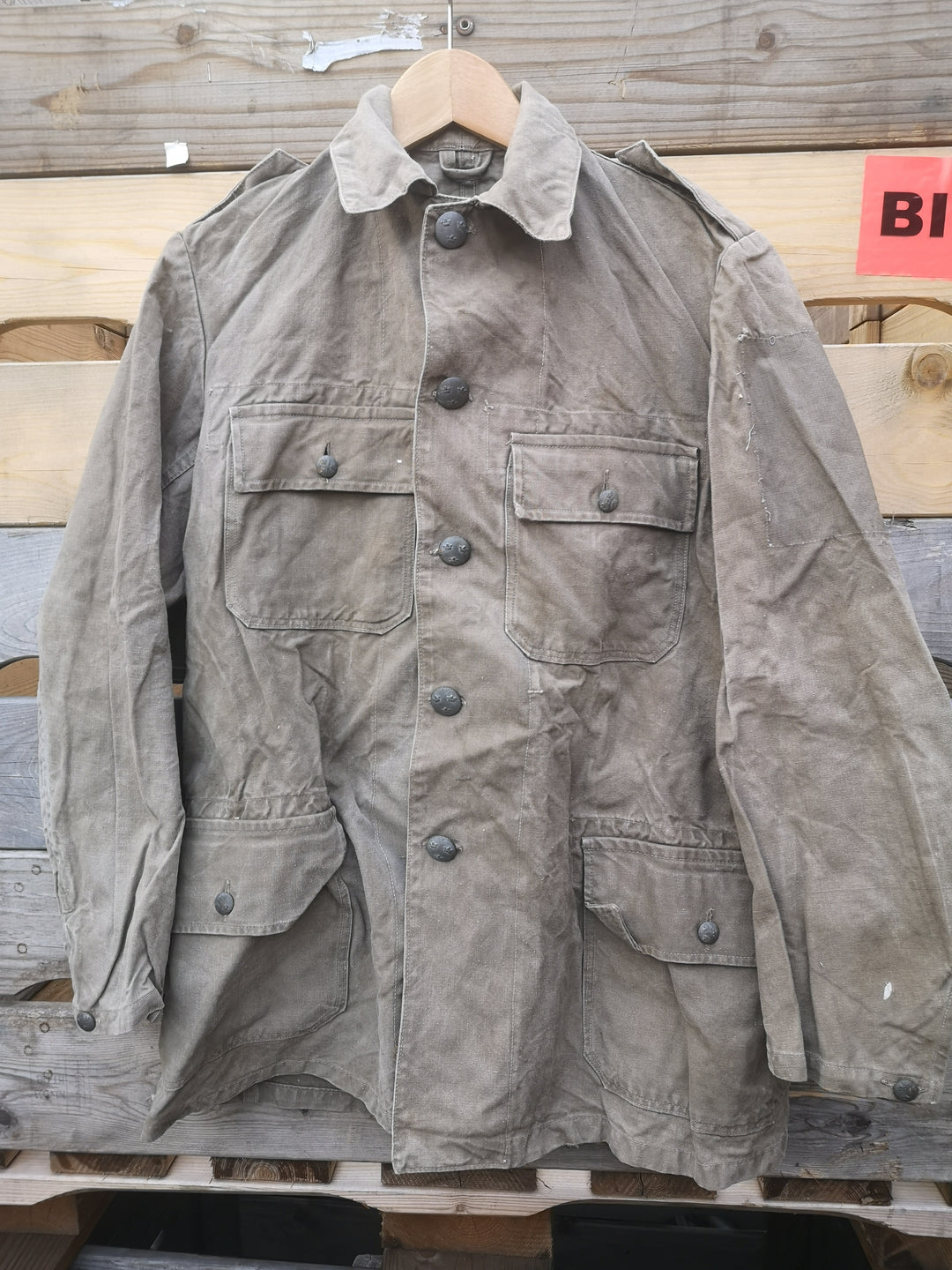 Vintage Khaki green army jacket with grey metal buttons, hanging from industrial wooden pallet.