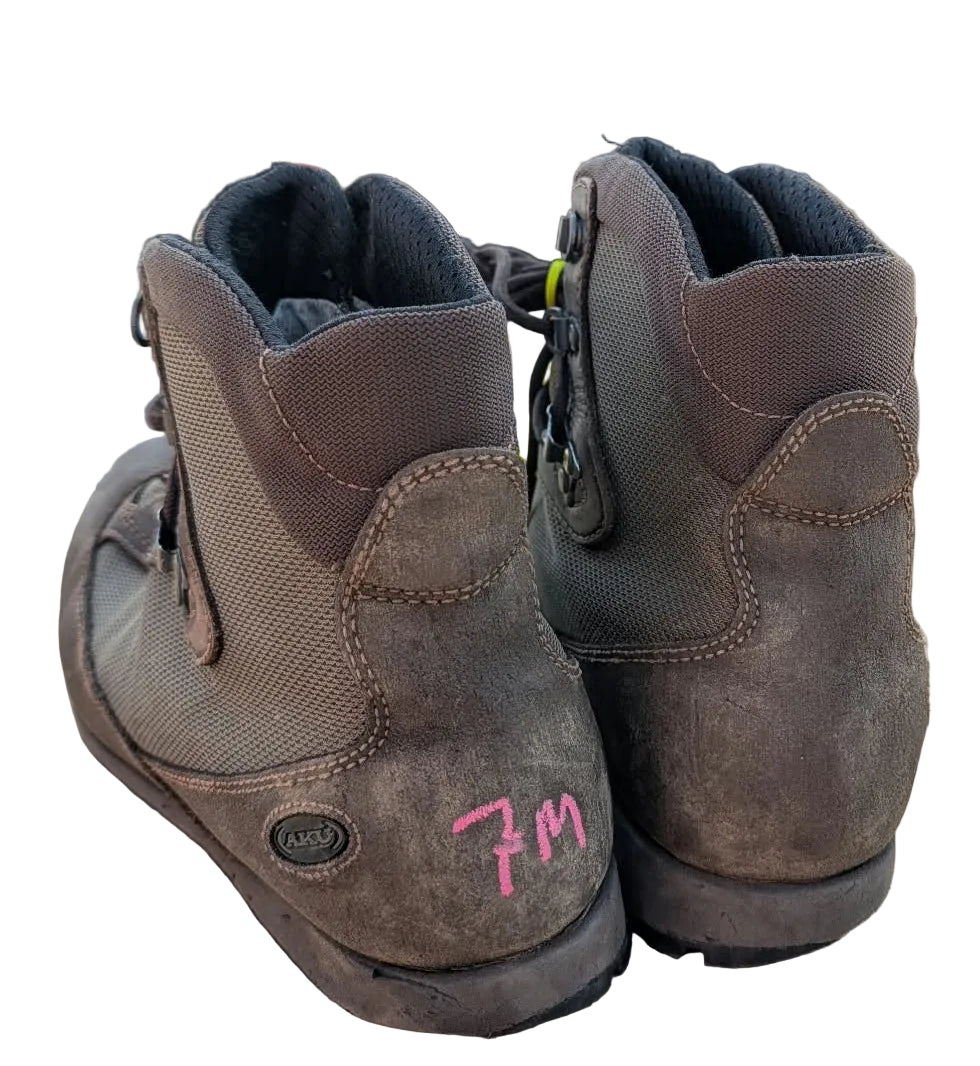 A pair of brown leather lace-up boots shown from the back, resting on a cardboard box in an outdoor or workshop setting with a wooden wall and a white van in the background.