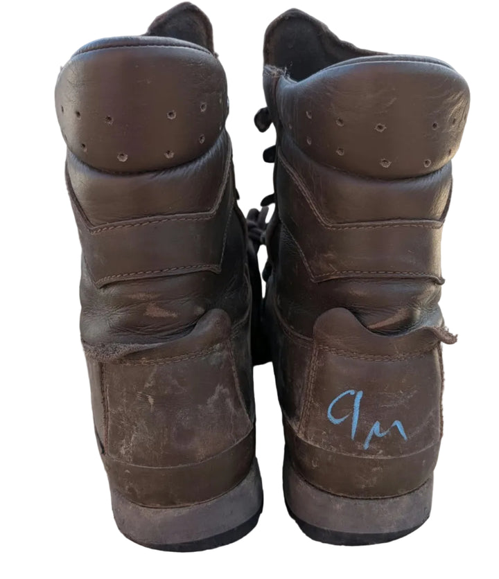 A pair of worn brown leather high-top boots standing upright on a cardboard box, photographed from the back. One boot has a blue handwritten marking near the heel.