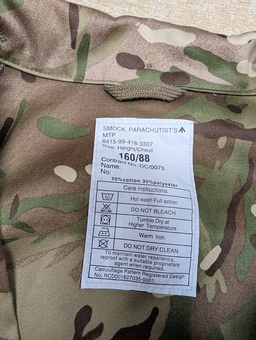 A white care label for a MTP 'Smock. Parachutist's' camouflage garment, laid flat on a camouflage-patterned fabric background.