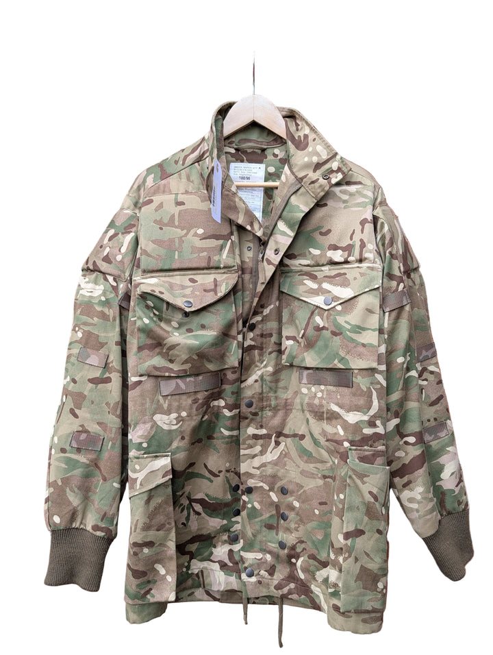 A military-style camouflage jacket hanging on a wooden hanger against a red brick wall.