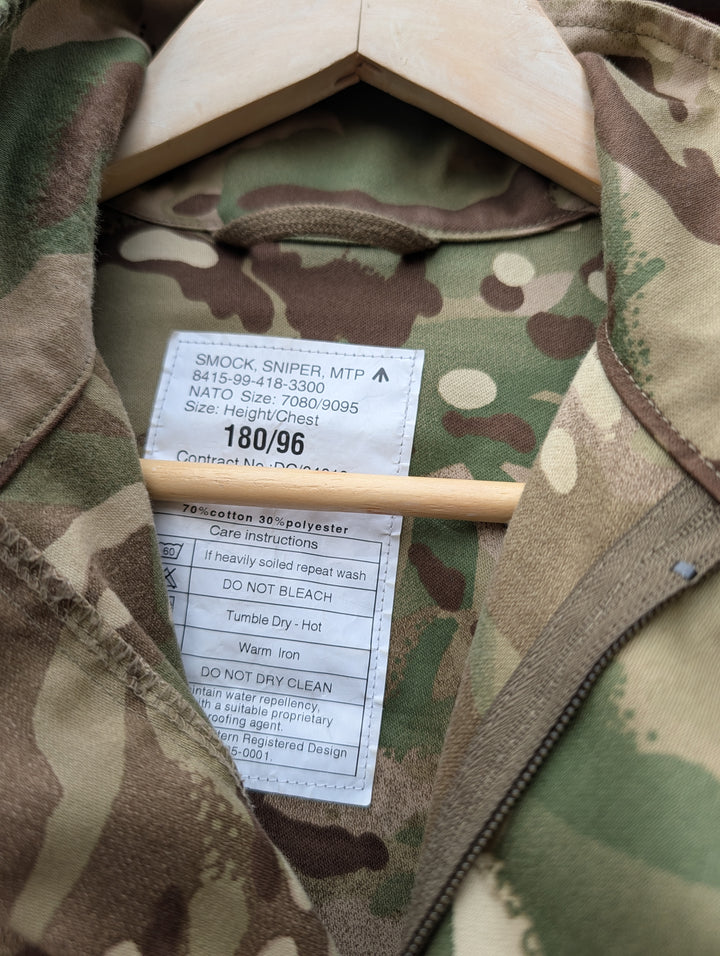 Close-up of a camouflage zip jacket on a wooden hanger, showing the interior collar, zipper, and a white sewn-in size/contract label.