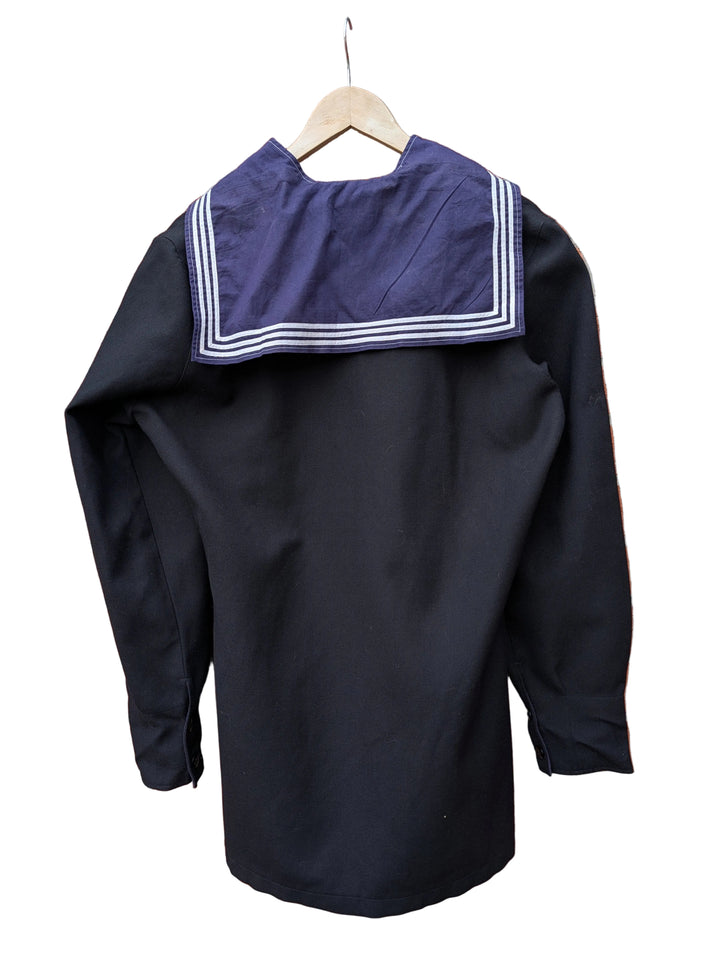 A black long-sleeve sailor-style top with a dark purple collar featuring white stripes, hanging on a wooden hanger against a red brick wall.