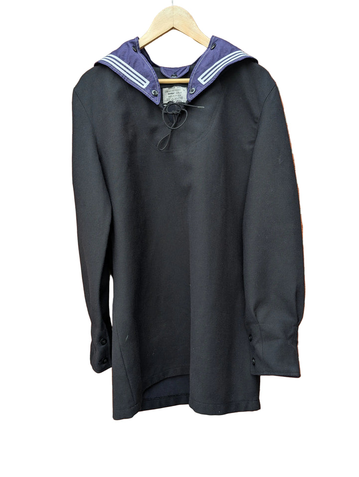 A black long-sleeve sailor-style top with a navy blue collar featuring white stripes, hanging on a wooden hanger against a red brick wall.