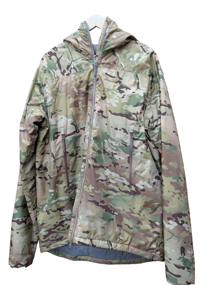 A hooded camouflage jacket with a full front zipper and zippered hand pockets, hanging on a white hanger against a red brick wall.