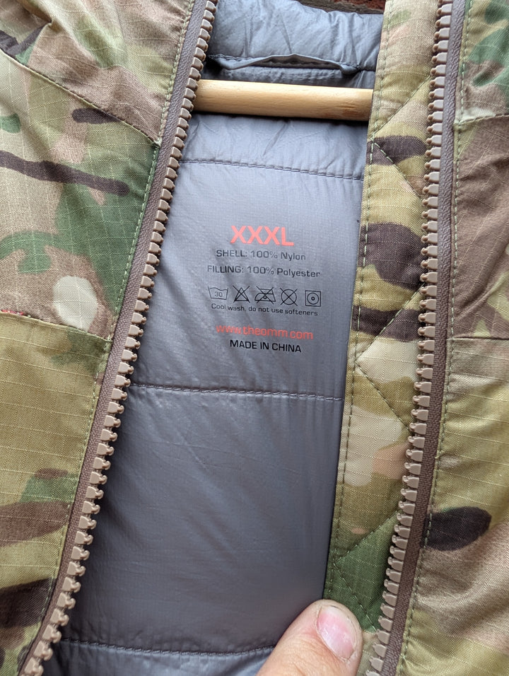 Close-up of the inside collar of a camouflage jacket with a gray padded lining, showing the size label 'XXXL' and a finger pointing at it.