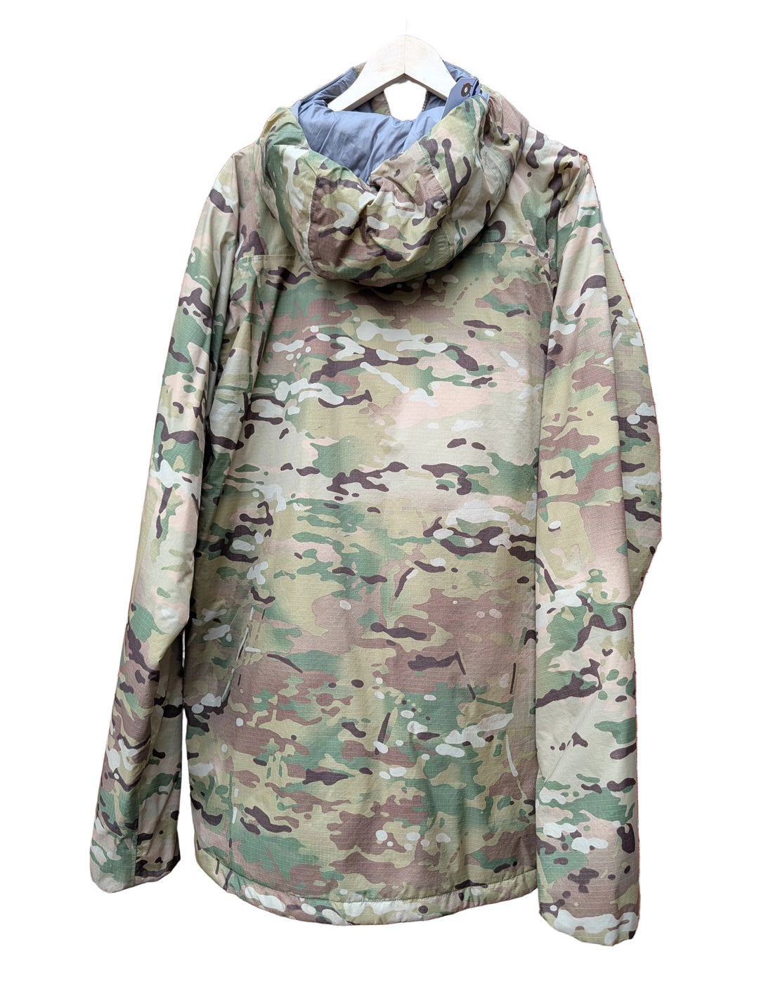 A hooded camouflage jacket hanging on a white hanger against a red brick wall. The jacket features a multi-terrain camouflage pattern in shades of green, brown, tan, and gray, with a light blue-gray lining visible inside the hood.