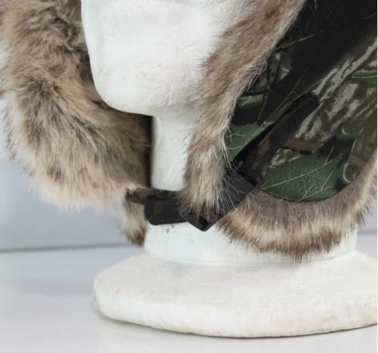 A close-up of a winter hat with thick brown and beige faux-fur trim and a green camouflage patterned side panel, displayed on a white mannequin head.