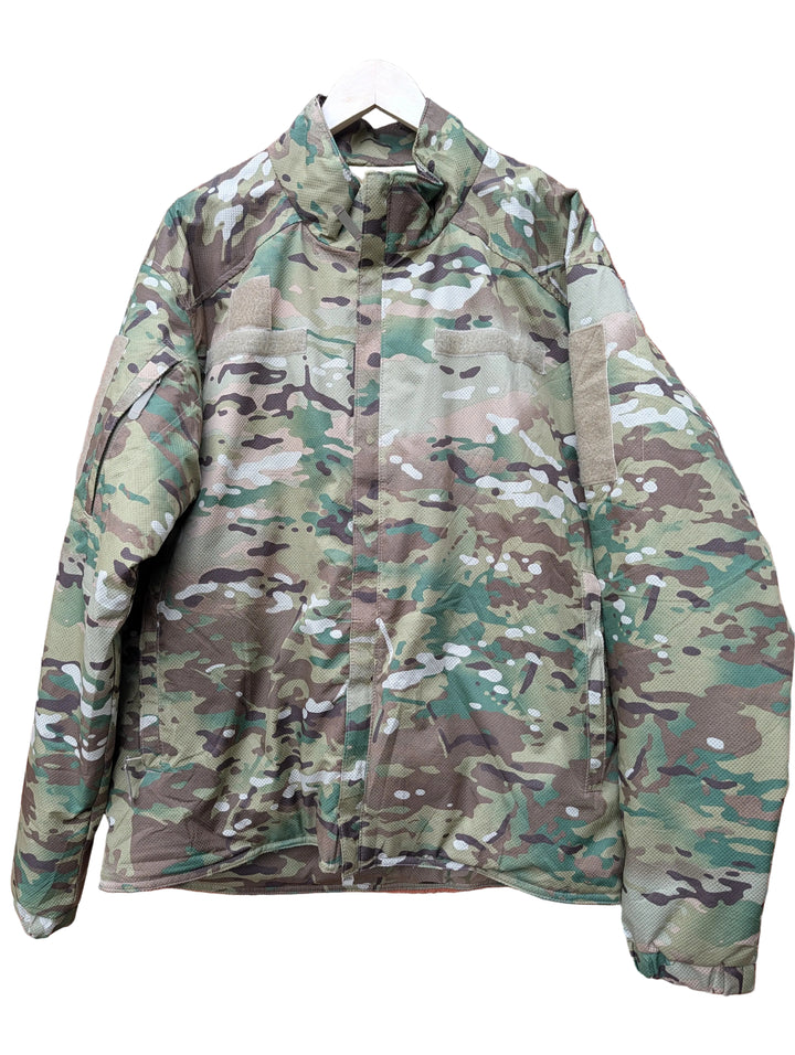 A camouflage military-style jacket hanging on a white hanger against a red brick wall.